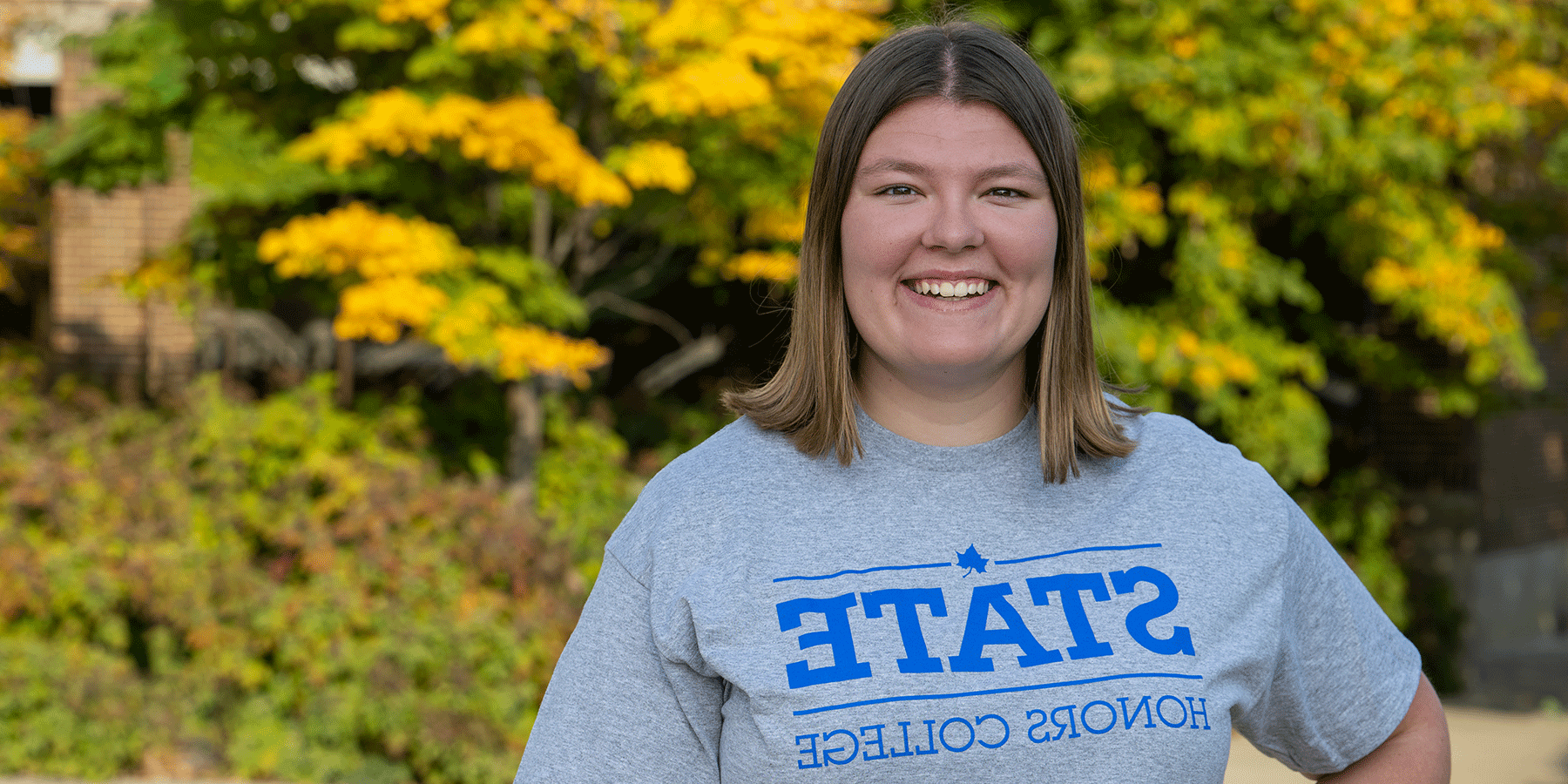 Honors student Kaylan Galey, smiling, with shoulder-length brown hair, is wearing a gray Indiana State Honors t-shirt. Trees with green and yellow leaves are visible in the background.